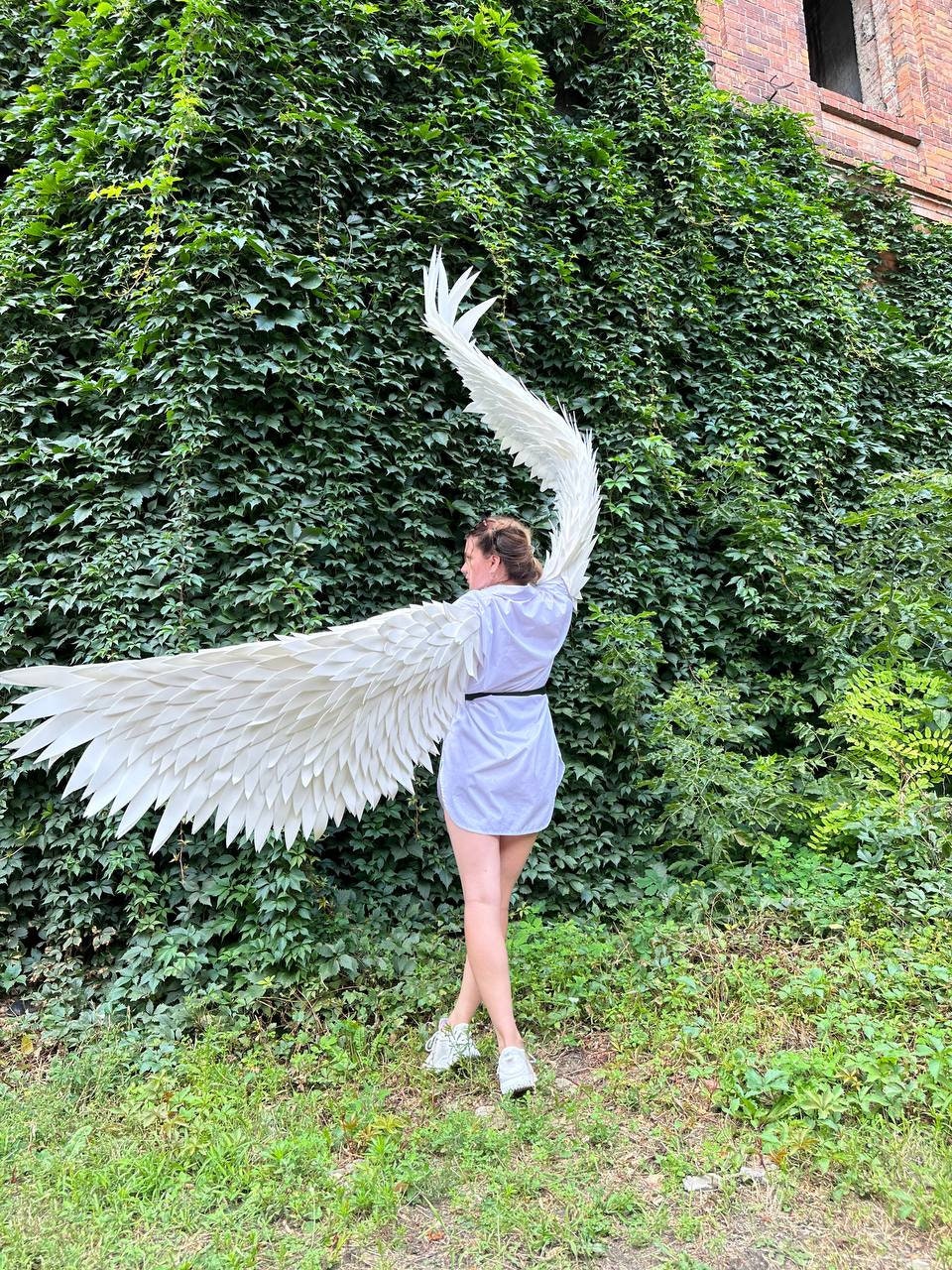 May include: A person wearing a white shirt and white shoes stands in front of a green ivy covered wall. They are holding large white angel wings.