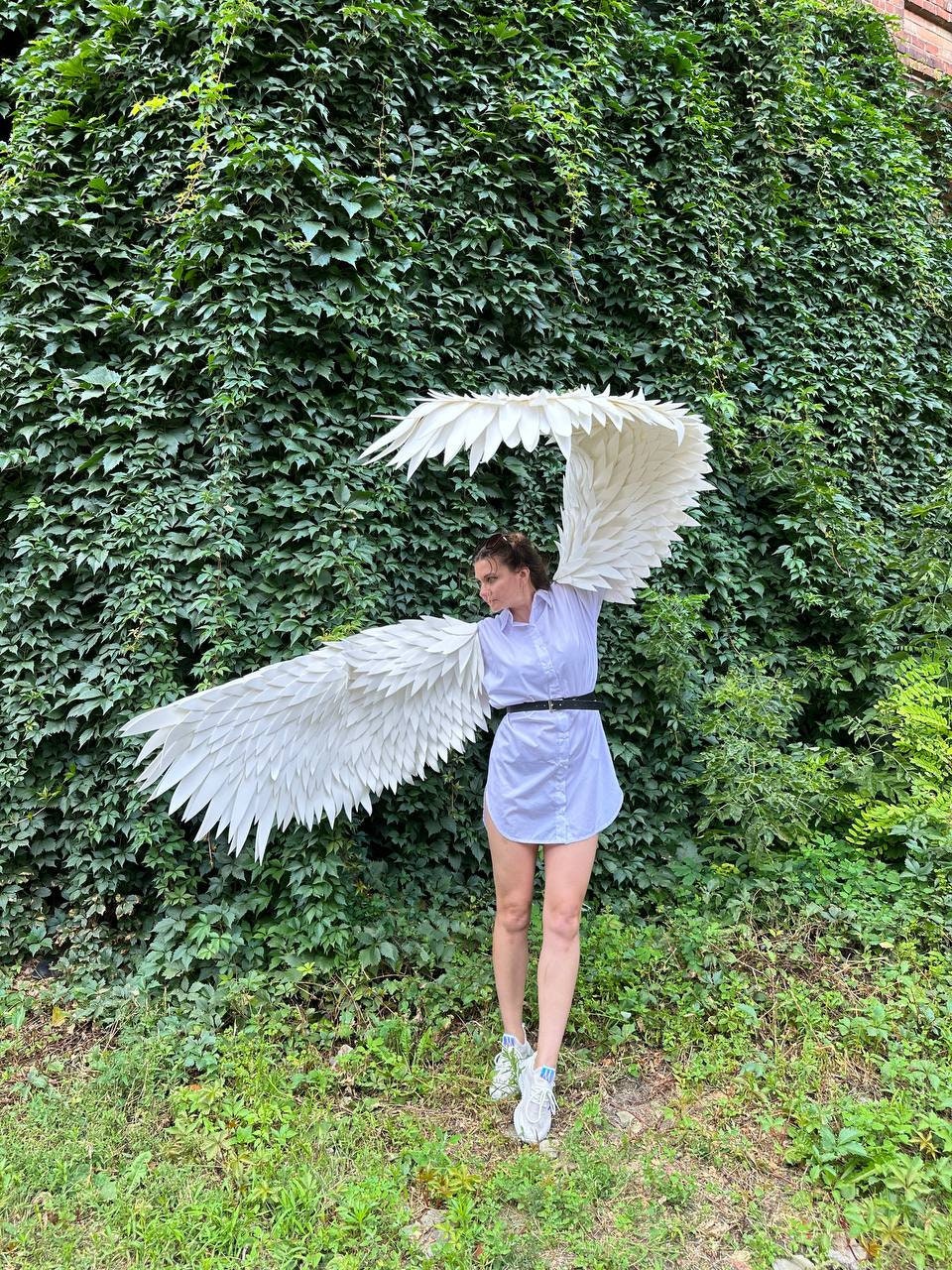 May include: A person wearing a white shirt and white sneakers is holding large white angel wings in front of a green ivy wall.