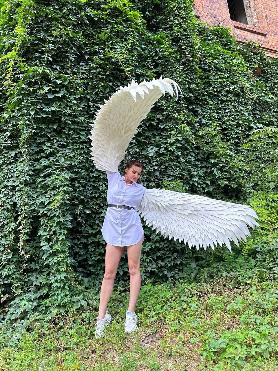 May include: A person wearing a white shirt and white sneakers is holding large white angel wings in front of a green ivy wall.