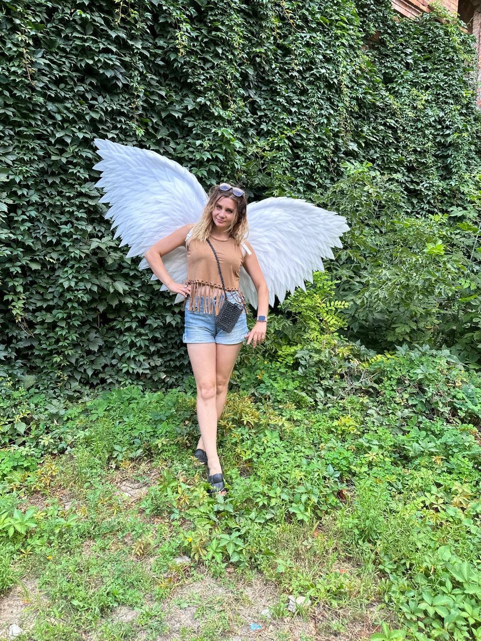 May include: A woman wearing a brown tank top and denim shorts poses with large white angel wings in front of a green ivy wall.