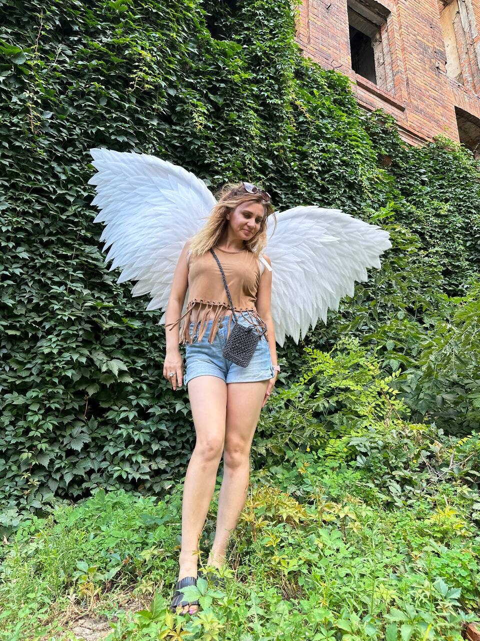 May include: A person wearing a brown tank top and denim shorts with white angel wings. The person is standing in front of a brick wall covered in green ivy.