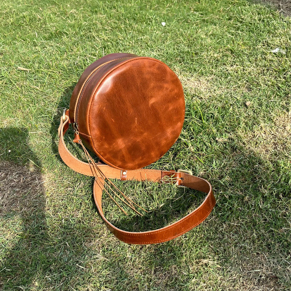 May include: A brown leather round crossbody bag with a long strap and a zipper closure.