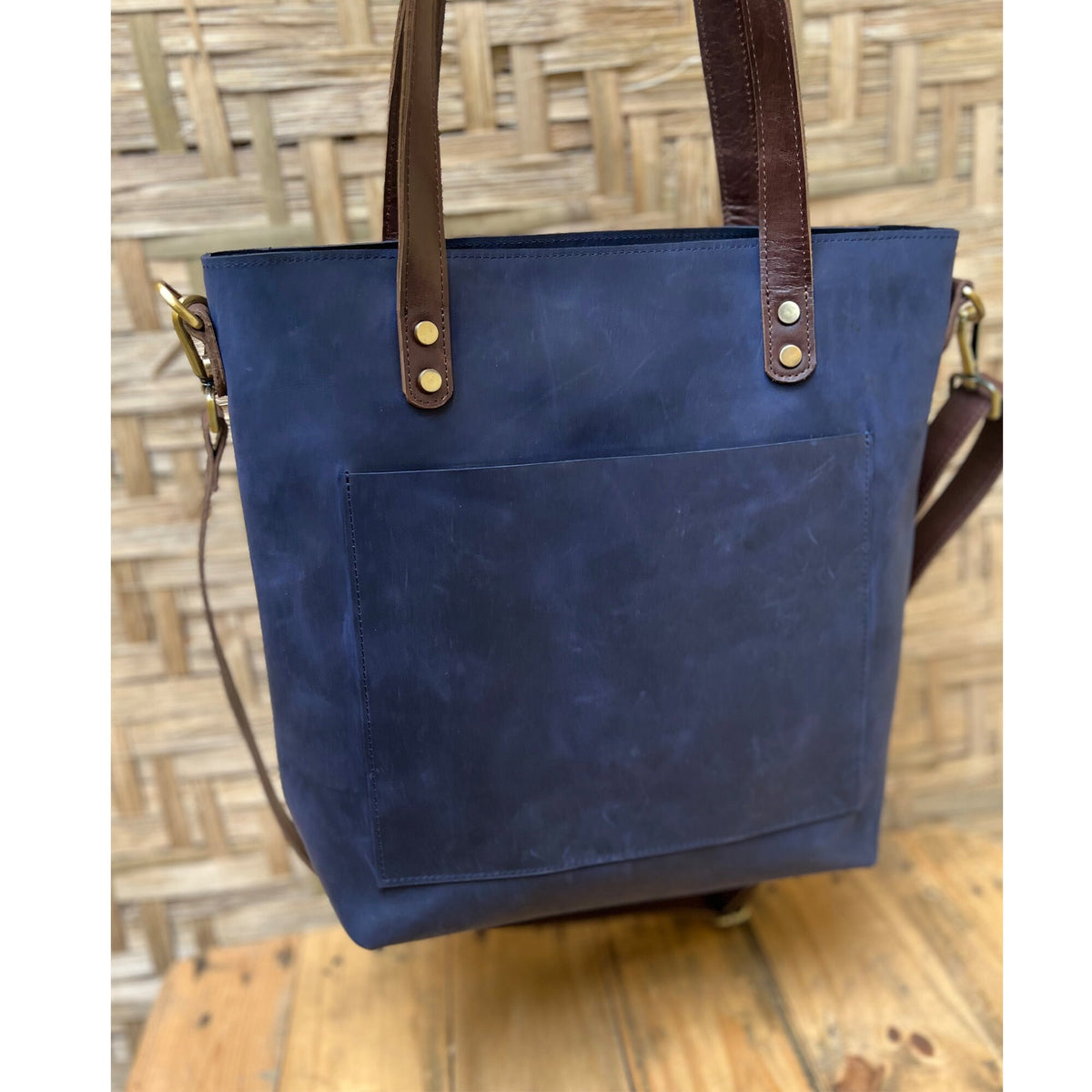 May include: A dark blue leather tote bag with brown leather straps and a front pocket. The bag has gold metal accents.