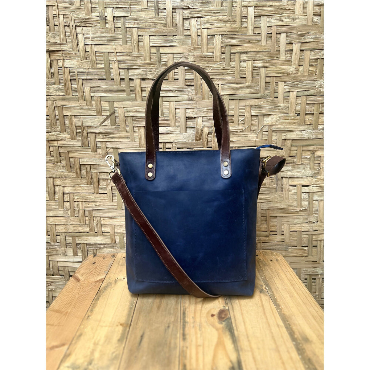 May include: A dark blue leather tote bag with brown leather straps and a zipper closure. The bag has a front pocket and a gold metal accent.