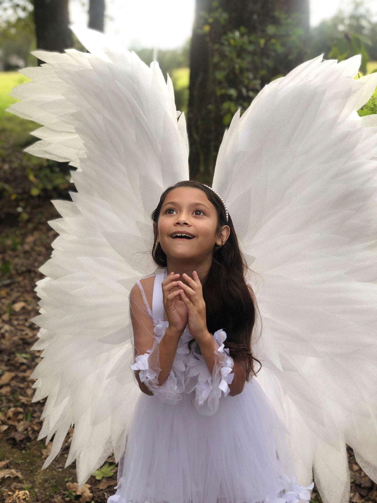 May include: A young girl wearing a white dress and angel wings, looking up with a smile. The wings are large and feathery, and the dress is decorated with lace.