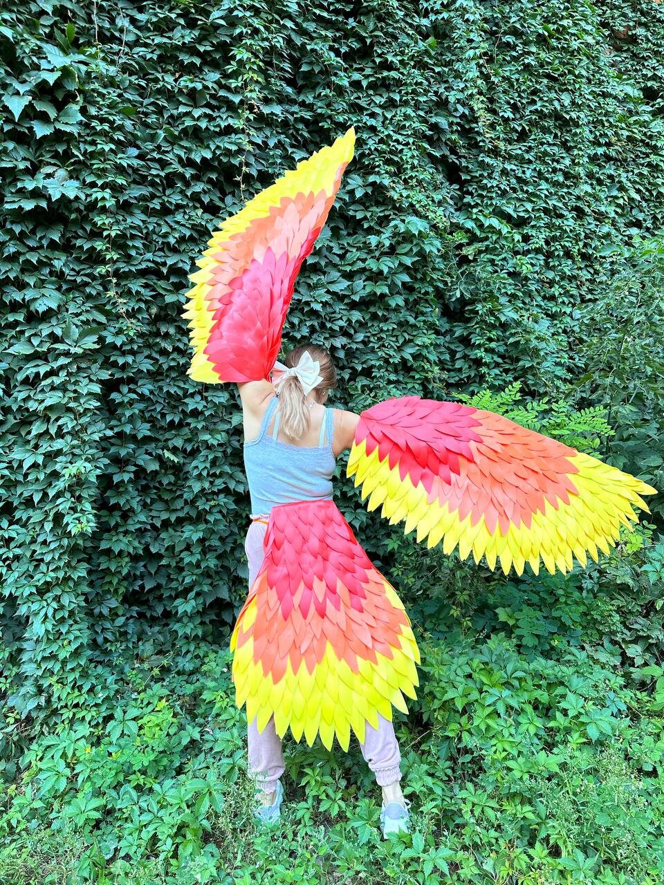 May include: A person wearing a costume with large, red and yellow wings. The wings are made of layered feathers and are attached to the person's back. The person is standing in front of a green leafy wall.