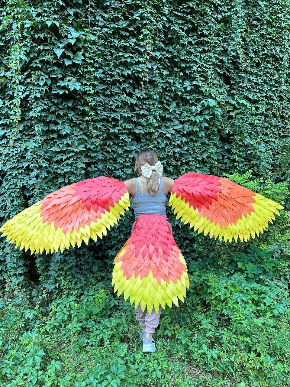 May include: A person wearing a colorful phoenix costume with large, layered wings and a matching skirt. The wings and skirt feature a gradient of red, orange, and yellow, resembling feathers. The background is a wall of green foliage.