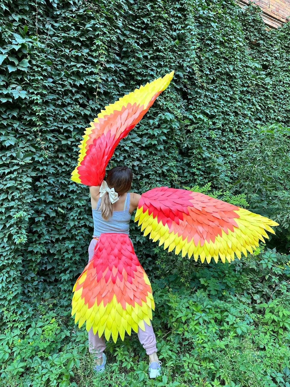 May include: A person wearing a costume with large, red and yellow wings. The wings are made of feathers and are attached to the person's back. The person is standing in front of a green wall.