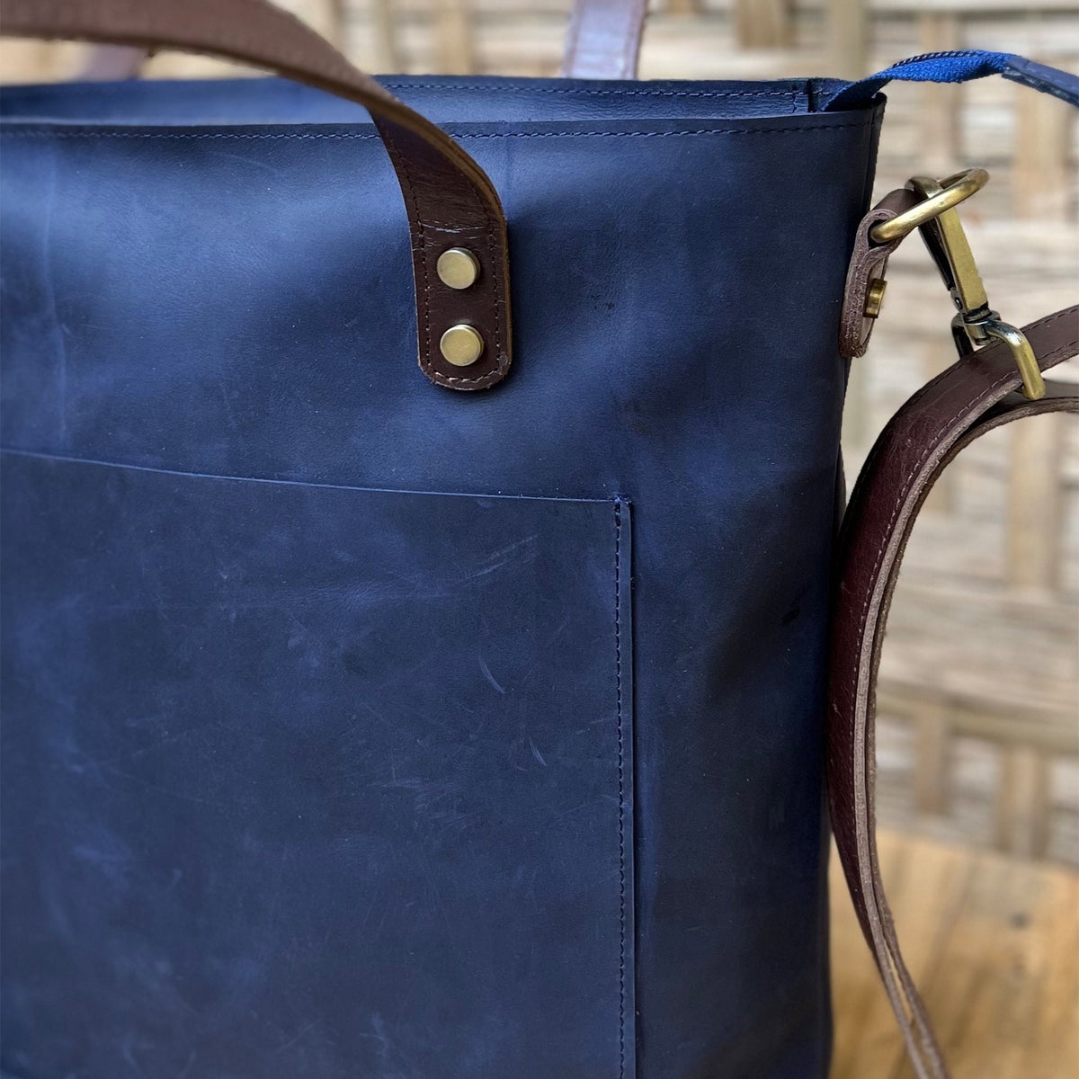 May include: A dark blue leather tote bag with a brown leather strap and a front pocket. The bag has brass hardware and a top handle.