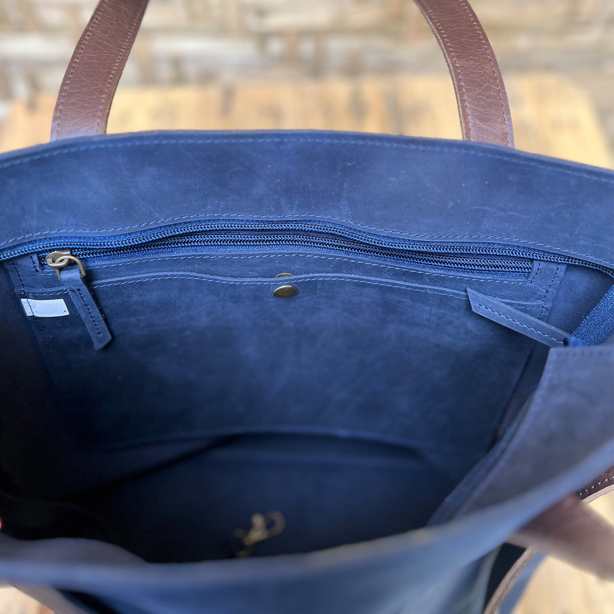 May include: Inside view of a navy blue leather tote bag with a brown leather handle. The bag has a zippered pocket and a small pocket with a snap closure.