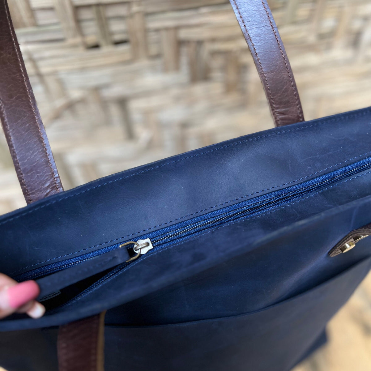 May include: A navy blue tote bag with brown leather handles and a zipper closure. The bag has a large pocket on the front.