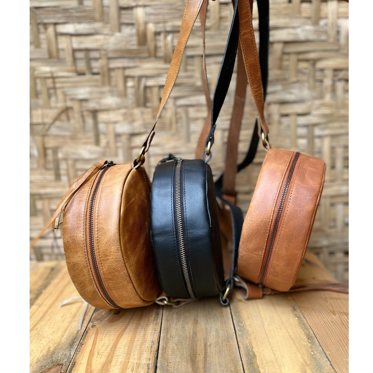 May include: Three round leather crossbody bags. One is brown, one is black, and one is a lighter brown. All three bags have a zipper closure and a long strap.