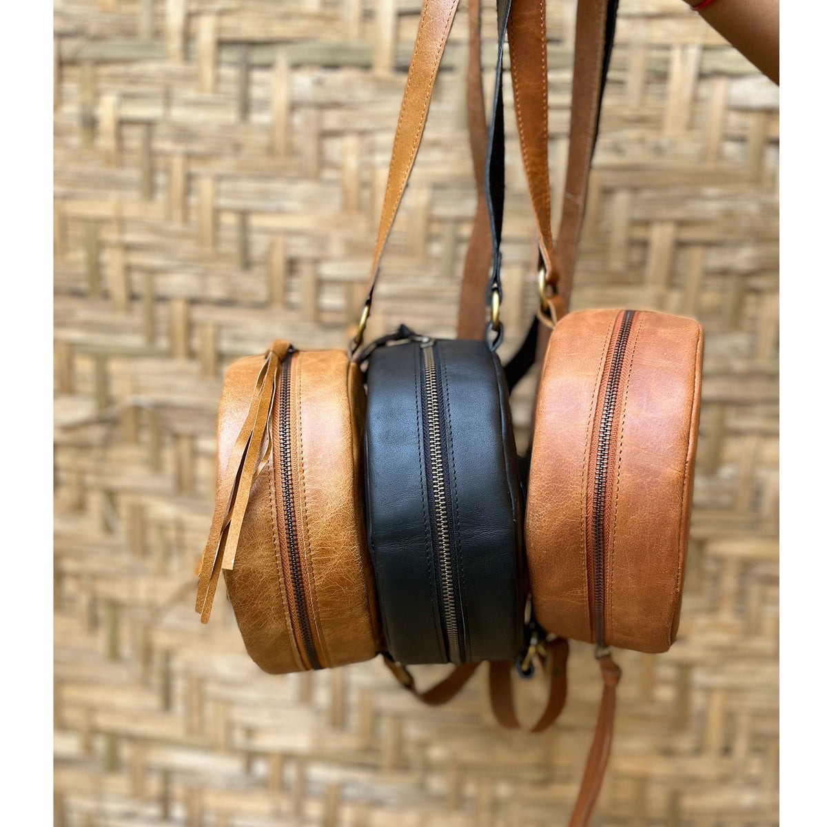 May include: Three round leather purses hanging from their straps. The purses are brown, black, and tan. The purses have zippers and are made of a soft, supple leather.