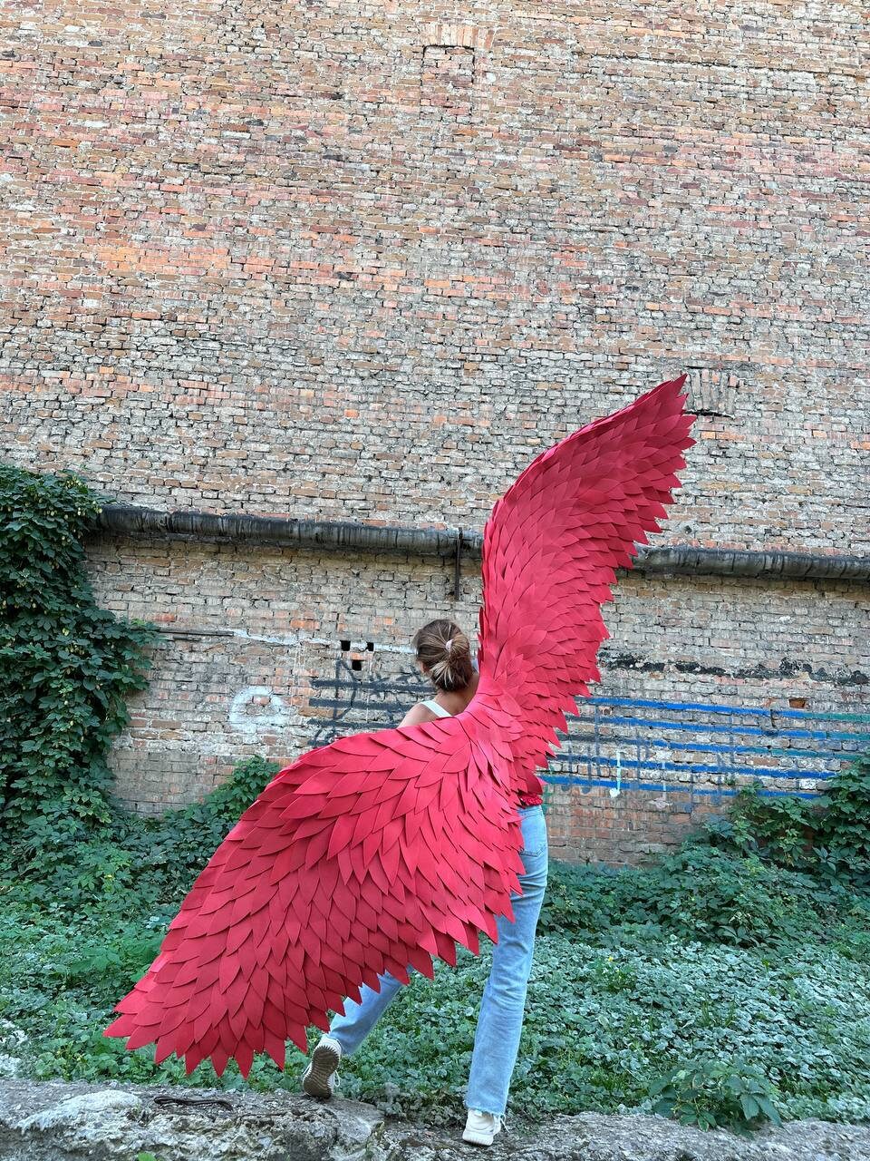 May include: A person wearing blue jeans and a white shirt is holding large, red, feathered wings behind their back. The wings are made of many individual feathers and are spread out wide.