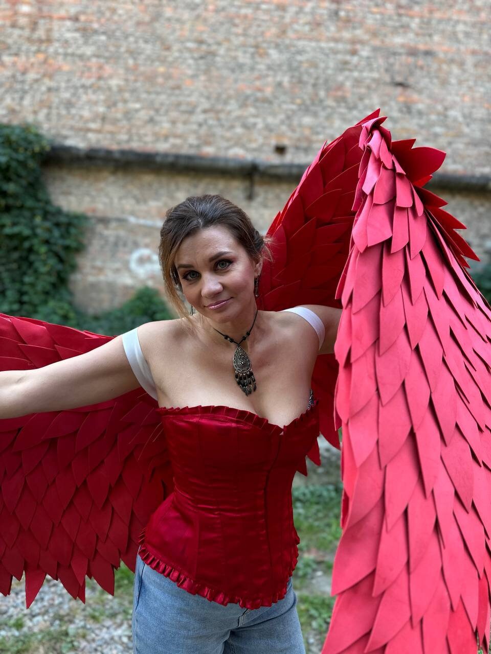 May include: A woman wearing a red corset and blue jeans with large red feathered wings.