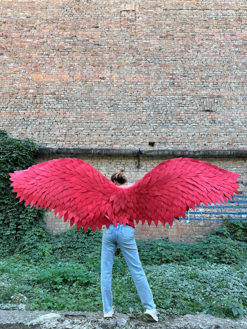 May include: A person wearing a pair of blue jeans and a red feathered angel wings costume. The wings are large and cover the person's back.