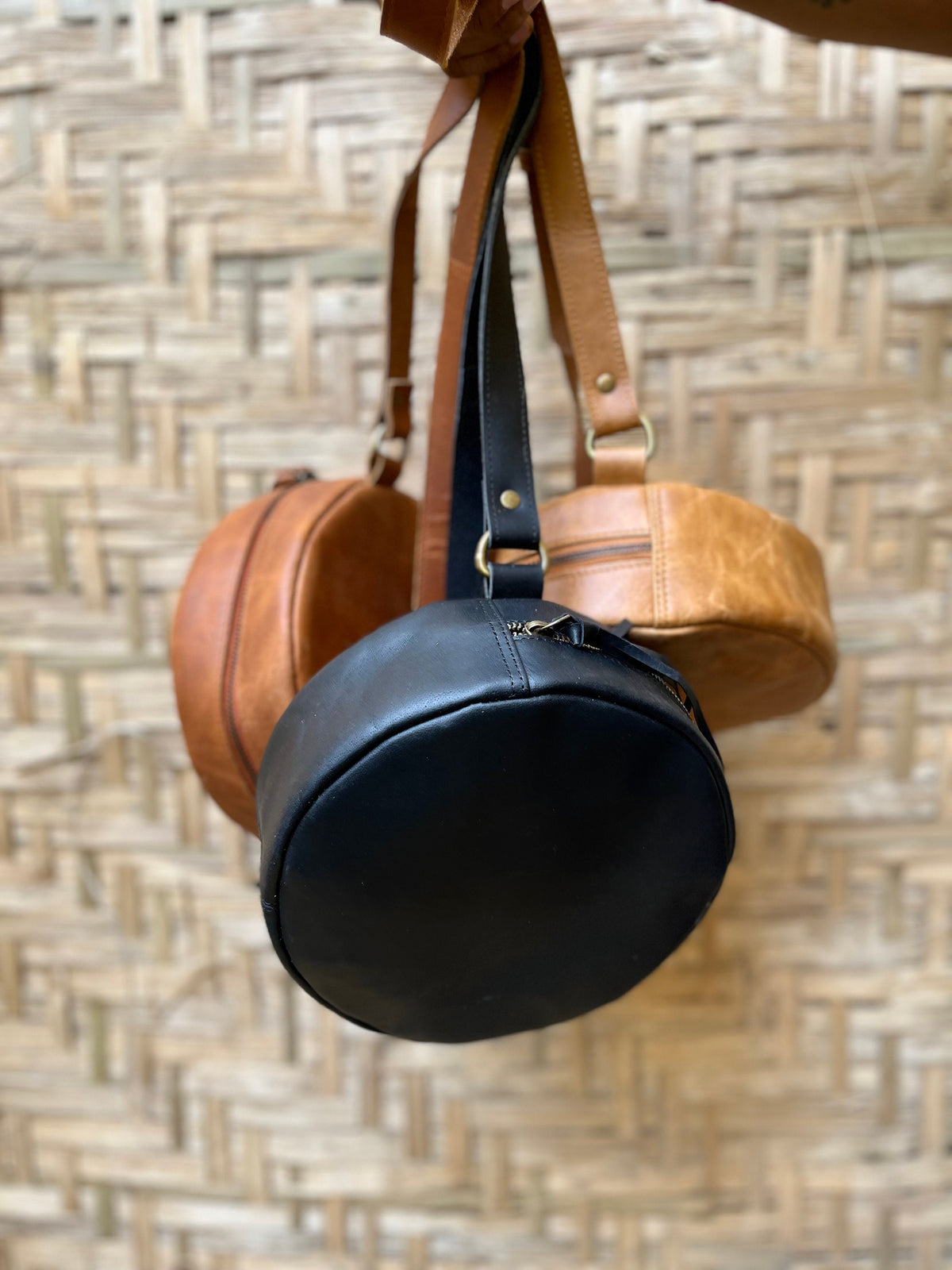 May include: Three round leather bags hanging from a woven wall. The top bag is brown leather, the middle bag is black leather, and the bottom bag is brown leather.