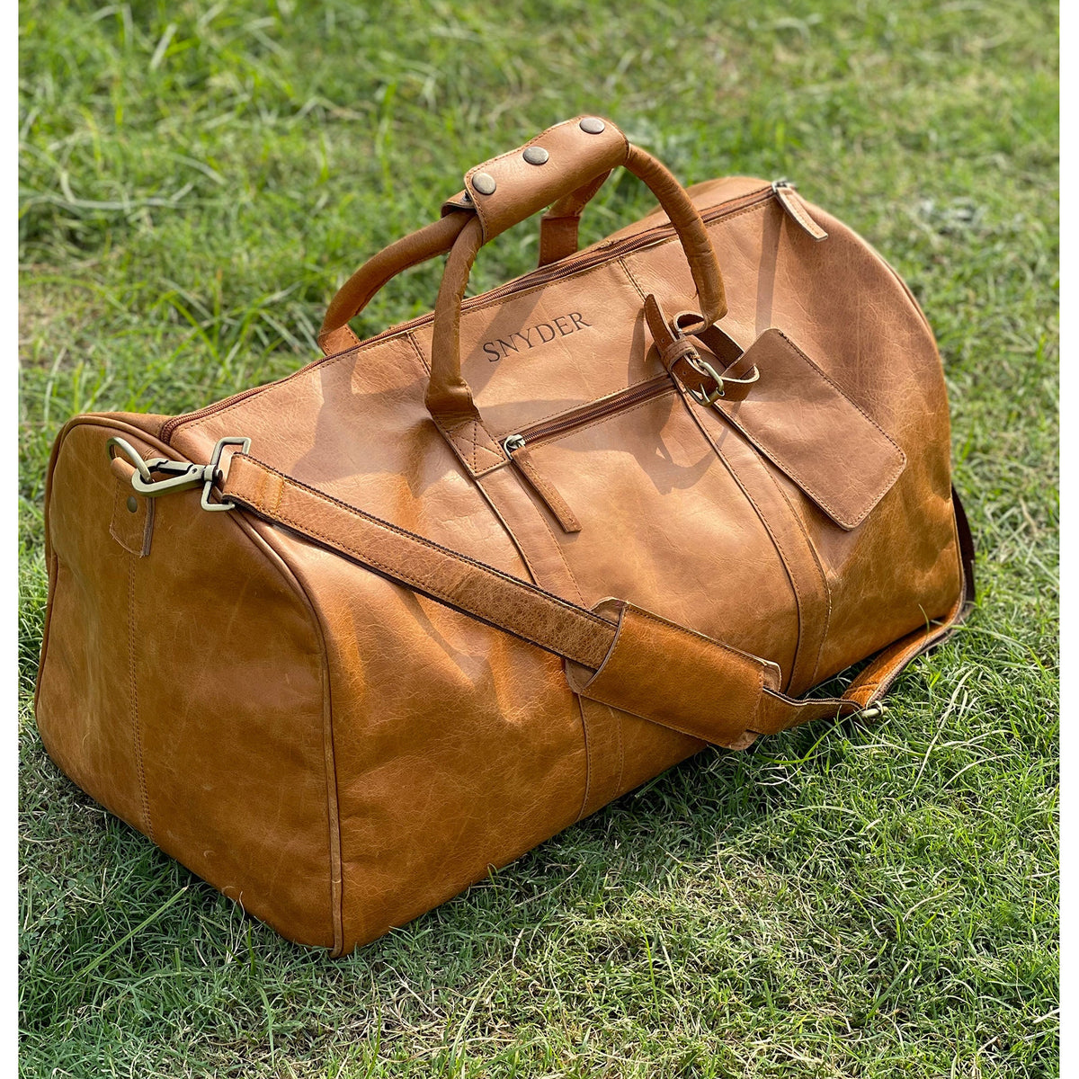 May include: A brown leather duffel bag with a top handle and a shoulder strap. The bag has a zipper closure and a small pocket on the front. The bag is personalized with the name "SNYDER" in silver lettering.