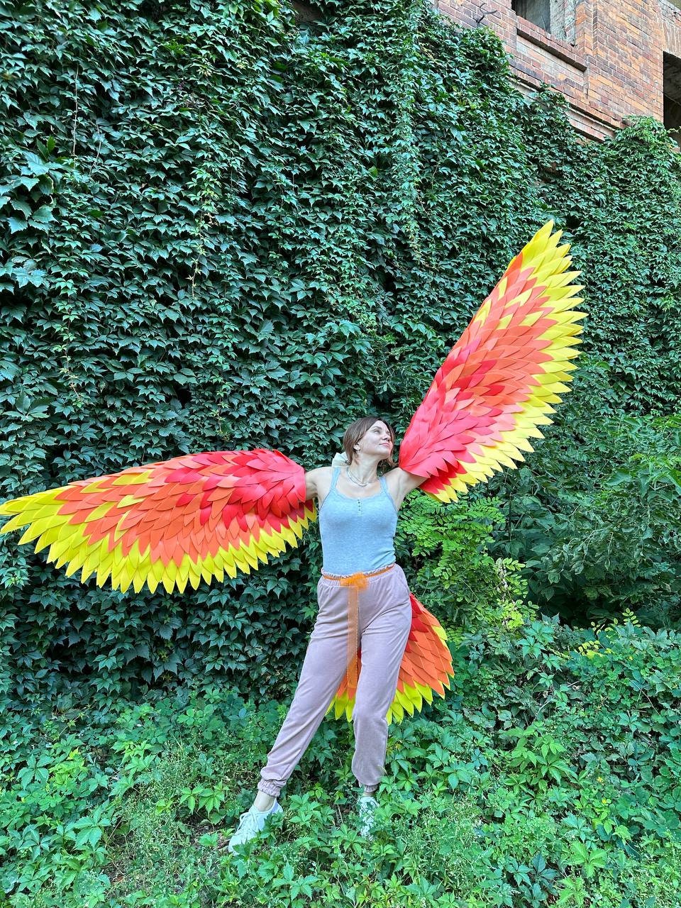 May include: A person wearing large, colorful phoenix wings with red, orange, and yellow hues, standing in front of a wall covered in green vines. The wings are spread wide, showcasing the vibrant colors and intricate design. The person is wearing a light blue tank top and pink pants.