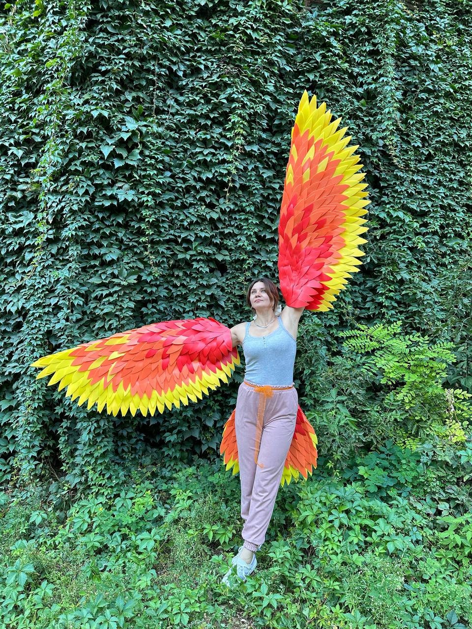 May include: A person wearing large, colorful wings in shades of red, orange, and yellow, resembling a bird's wings. The wings are spread wide against a backdrop of green foliage. The person is wearing a gray tank top and pink pants.