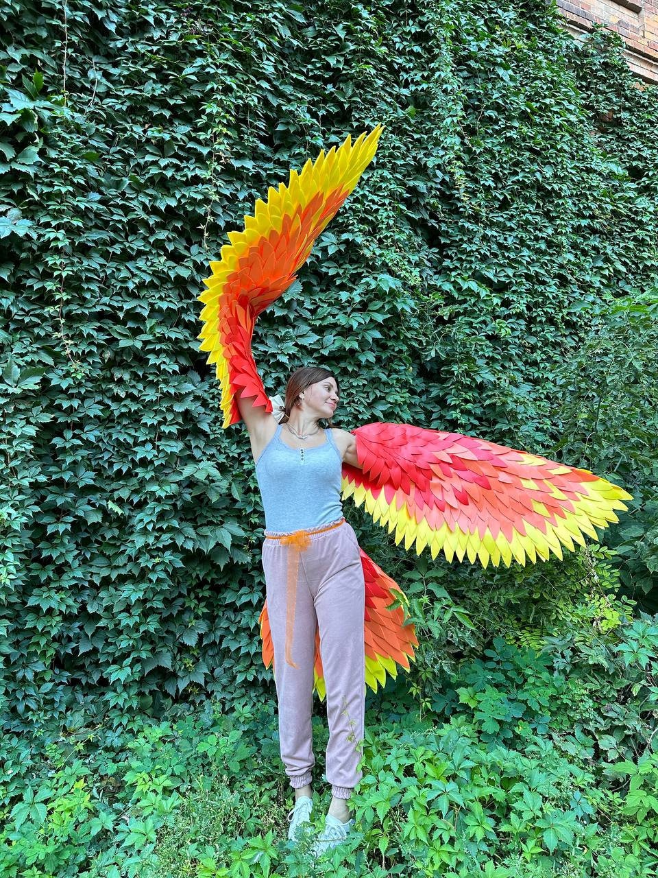 May include: A pair of vibrant, handmade phoenix wings in shades of red, orange, and yellow. The wings are made of layered, textured material, and are spread wide against a backdrop of lush green foliage. The wings are designed to be worn.