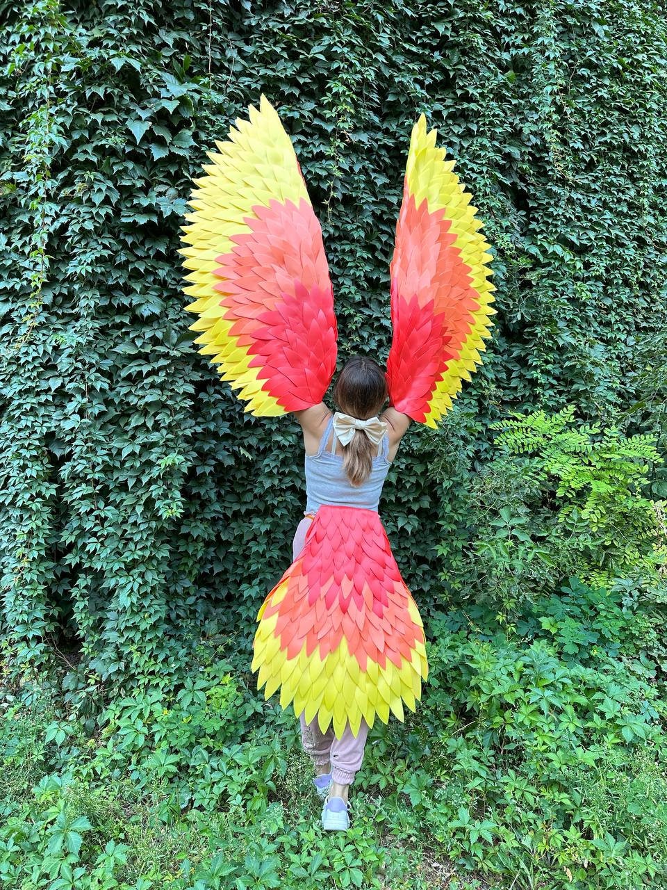 May include: A person wearing a costume with large, red and yellow wings. The wings are made of feathers and are attached to the person's back. The person is standing in front of a green leafy wall.