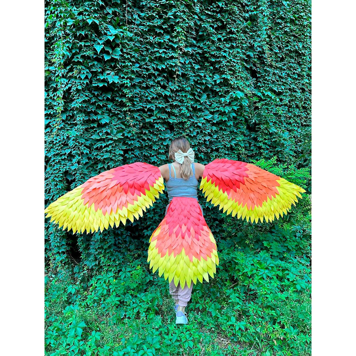May include: A person wearing a colorful costume with large, feathered wings and a matching skirt. The wings and skirt feature a gradient of red, orange, and yellow, resembling a bird. The background is a wall of green foliage.