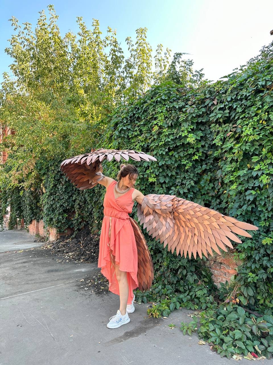 May include: A person wearing a coral-colored dress and large, brown, layered wing costume. The wings are outstretched, and the person is standing on a sidewalk in front of a green ivy-covered wall.