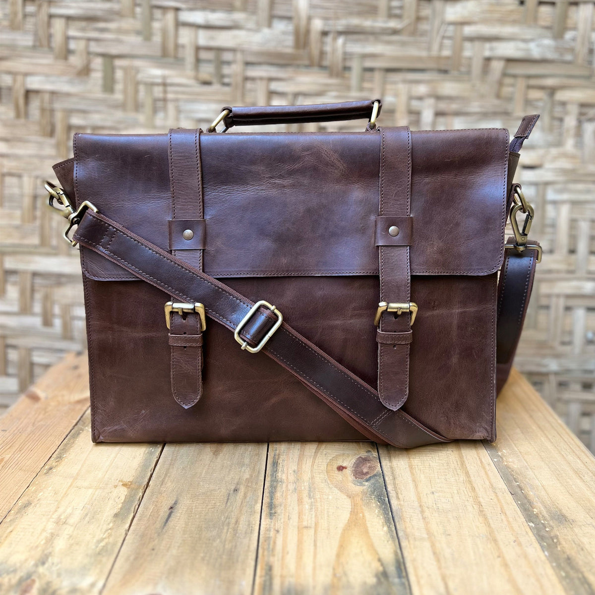 May include: A brown leather briefcase with a long adjustable strap and two buckles on the front. The briefcase has a top handle and a flap closure.
