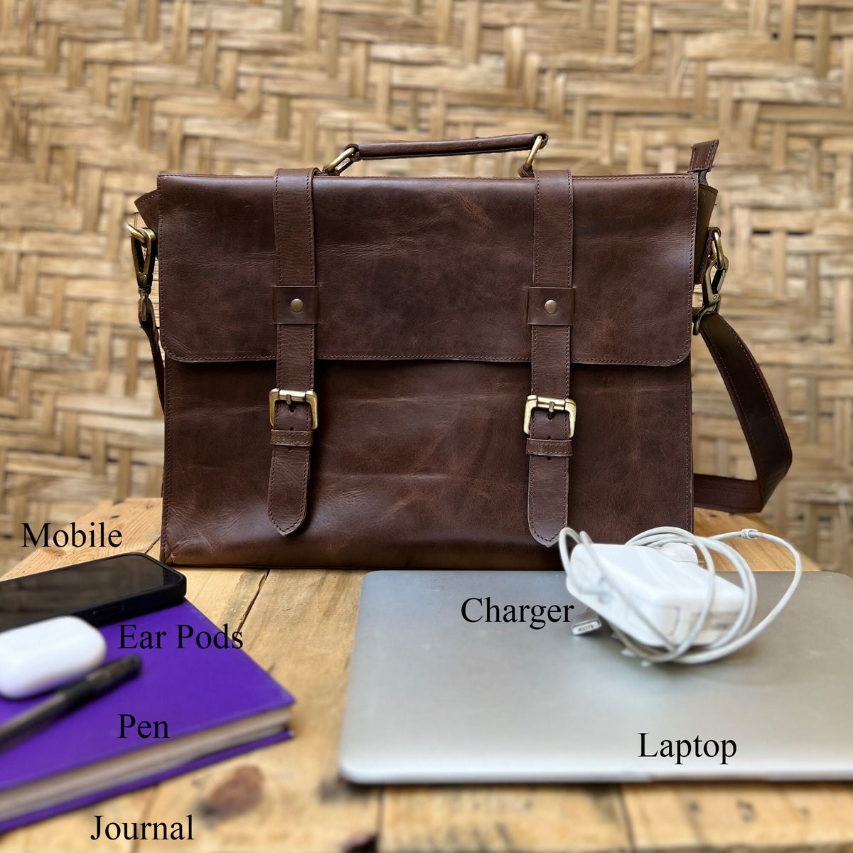 May include: A brown leather briefcase with two buckles on the front. The briefcase is sitting on a wooden table with a laptop, charger, phone, journal, pen, and ear pods.