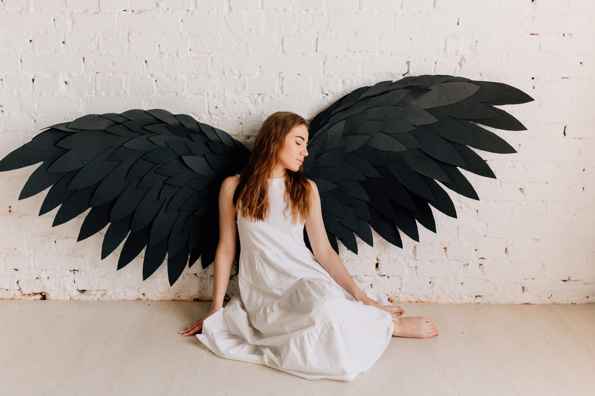 May include: A person wearing a white dress sits in front of a white brick wall with large black paper wings behind them.