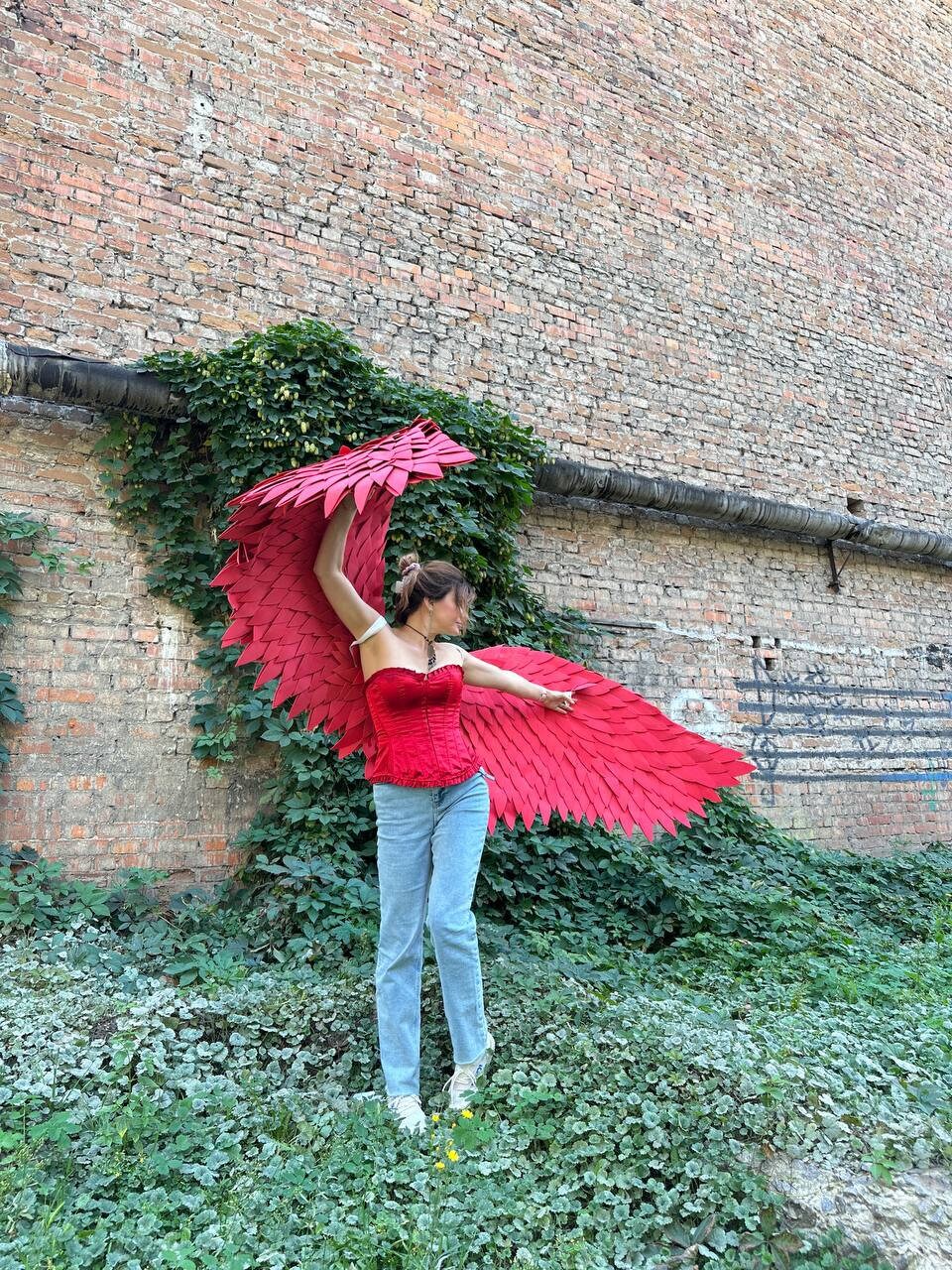 May include: A person wearing a red corset and blue jeans is holding large red wings made of paper or fabric. The person is standing in front of a brick wall with green vines growing on it.
