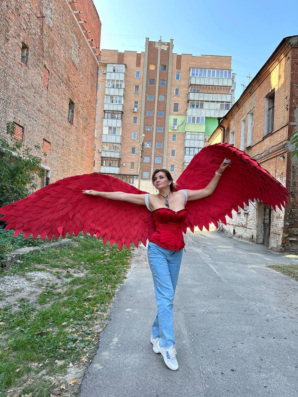 May include: A person wearing a red corset and blue jeans with large red feathered wings spread out behind them. The person is standing in a narrow alleyway between two brick buildings.