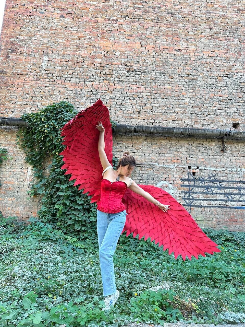 May include: A person wearing a red corset and blue jeans poses with large red wings made of paper or fabric. The wings are spread wide and the person is looking up.