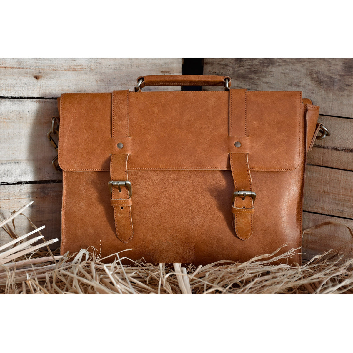May include: A brown leather briefcase with two buckles and a strap. The briefcase is sitting on a bed of straw.