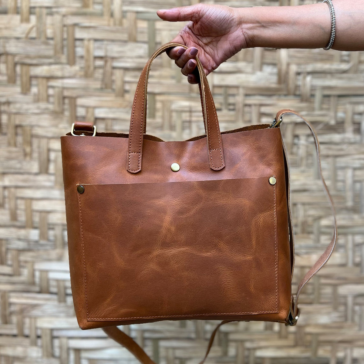 May include: A brown leather tote bag with a top handle and a long shoulder strap. The bag has a large front pocket and a brass button closure.