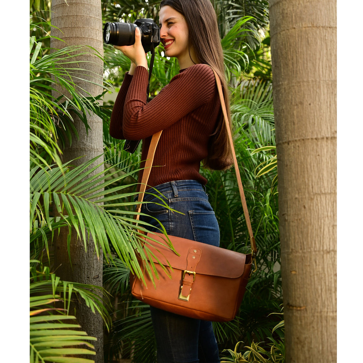 May include: A woman wearing a maroon sweater and blue jeans is holding a camera and wearing a brown leather camera bag with a strap over her shoulder.