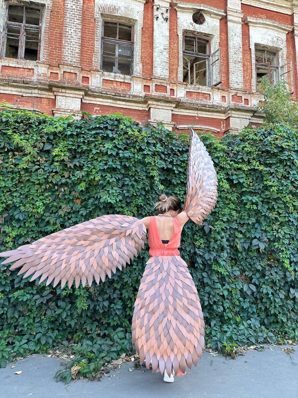 May include: A person wearing a costume with large, feathered wings and a matching skirt. The wings are a mix of brown and tan, and the dress is coral. The background features a brick building and green foliage.
