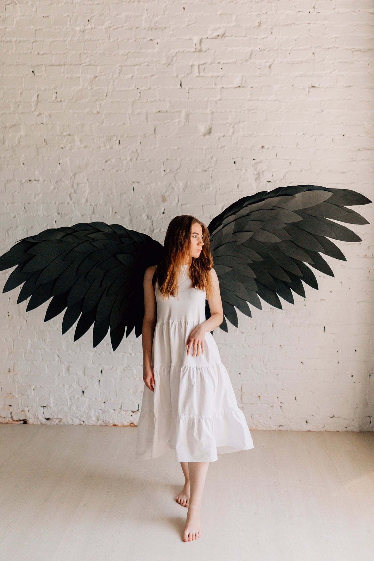 May include: A woman wearing a white dress stands in front of a white brick wall with large black paper wings behind her.