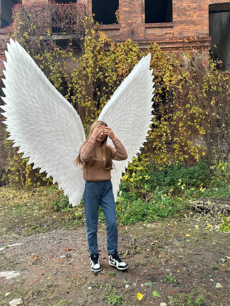 May include: A person wearing a brown sweater and blue jeans stands in front of a brick building with ivy growing on it. They are wearing large white angel wings.