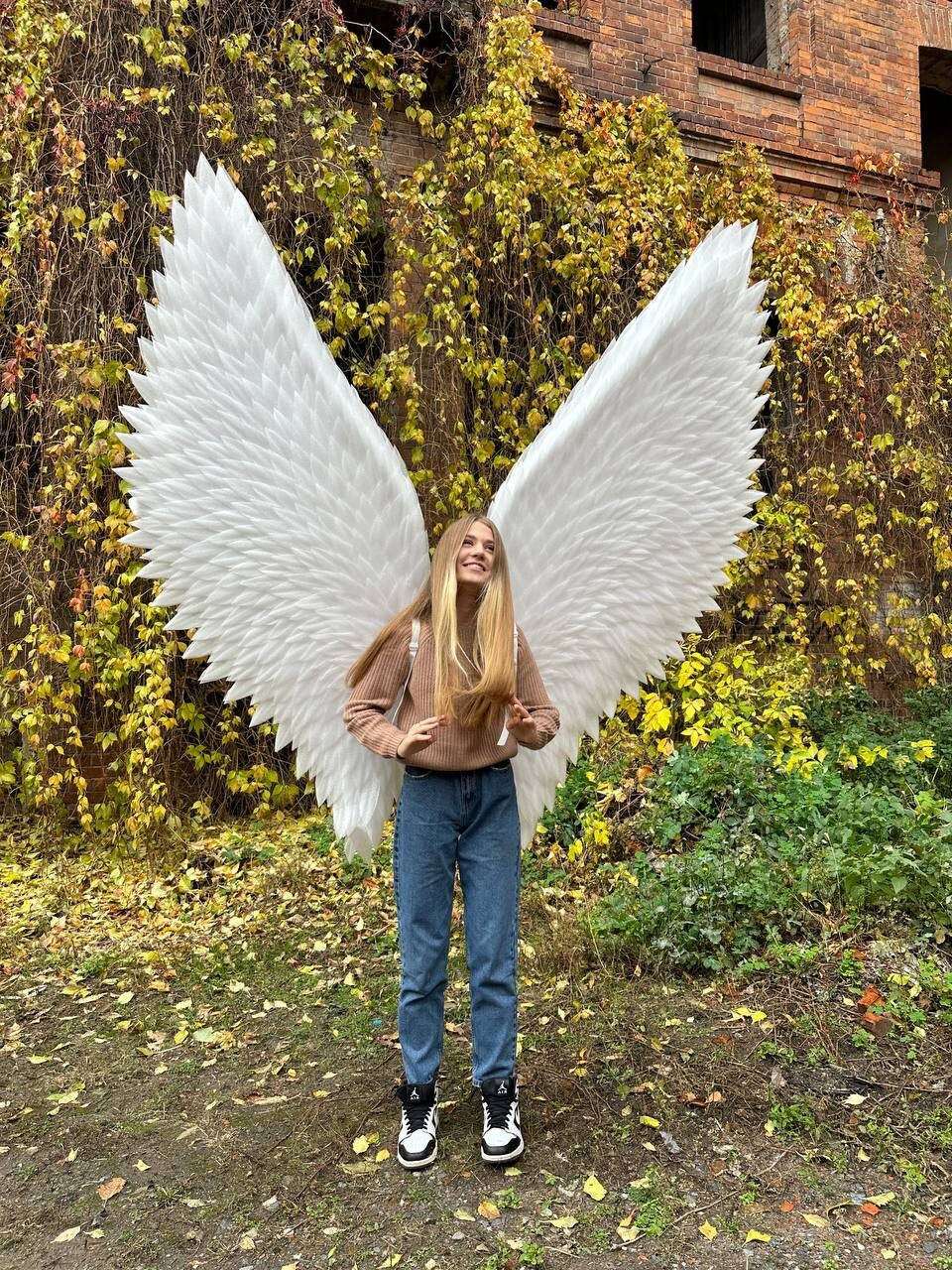 May include: A person wearing a brown sweater and blue jeans is standing in front of a brick wall covered in ivy. They are holding large white angel wings.