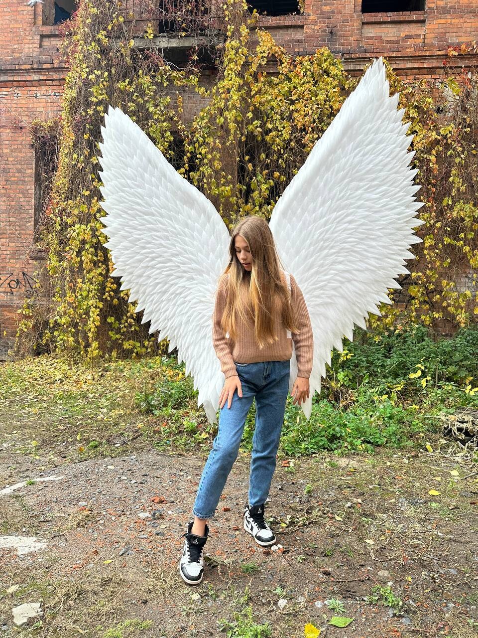 May include: A person wearing a brown sweater and blue jeans is standing in front of a brick wall with white angel wings attached to their back. The wings are large and feathery, and the person is looking down at the ground.