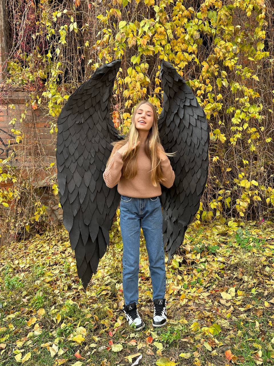 May include: A person wearing blue jeans and a brown sweater stands in front of a backdrop of yellow leaves. They are wearing black angel wings with a feathery texture.
