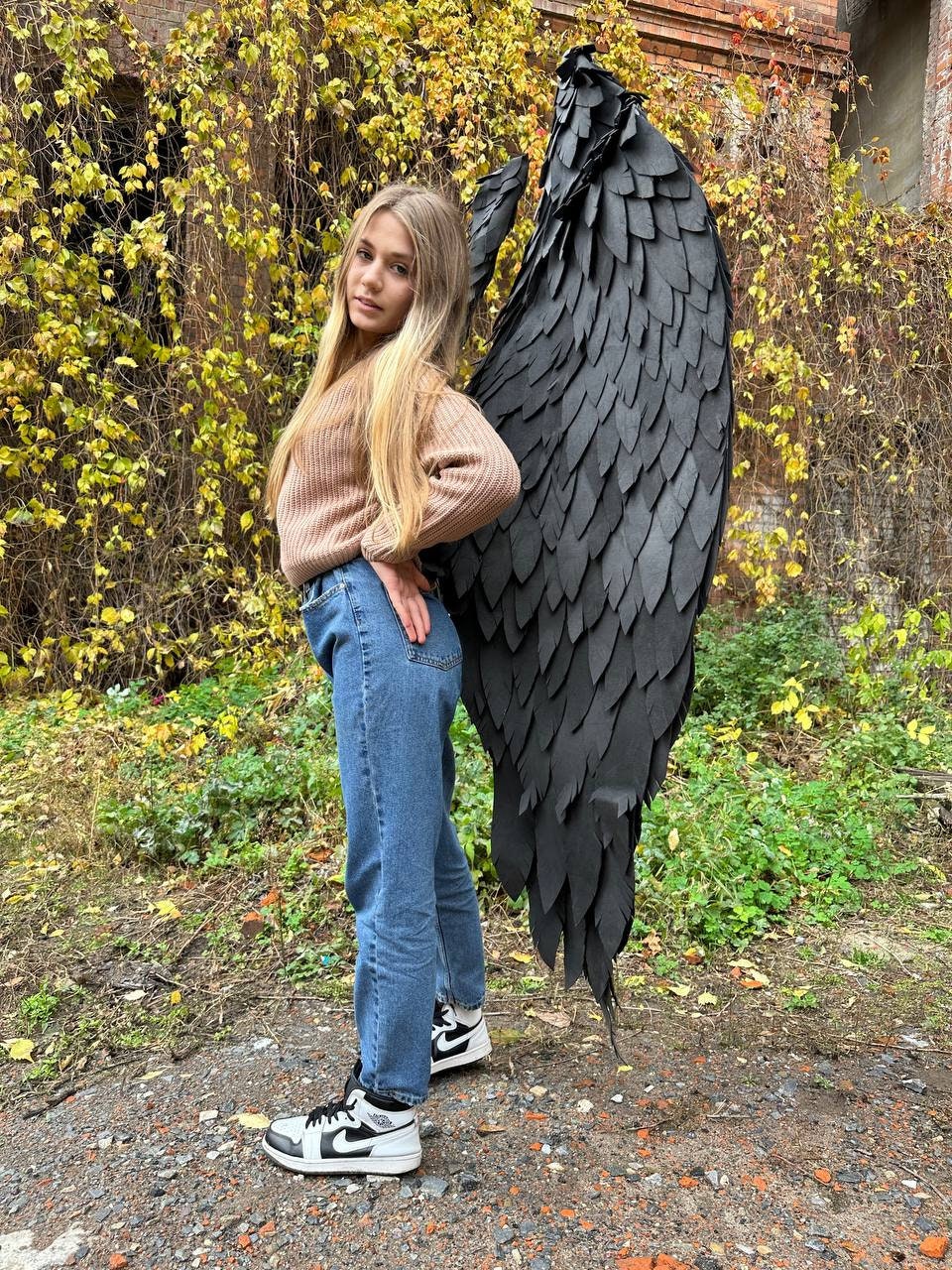 May include: A young person wearing blue jeans and a brown sweater stands with their back to the camera, holding large black feathered wings behind them.