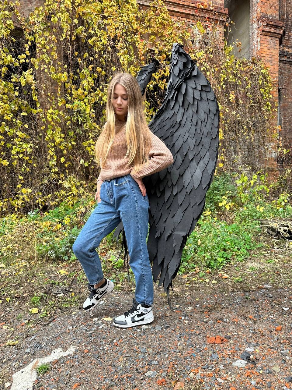 May include: A person wearing a brown sweater, blue jeans, and black and white sneakers, standing in front of a large pair of black angel wings.