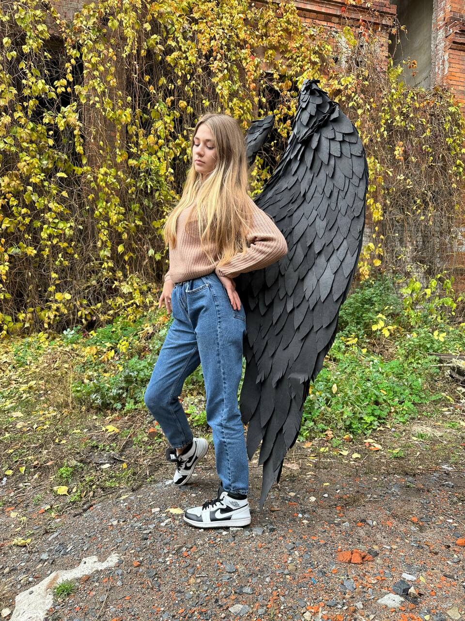 May include: A person wearing blue jeans and a brown sweater stands with large black feathered wings behind them. The wings are made of paper or fabric and are very detailed.