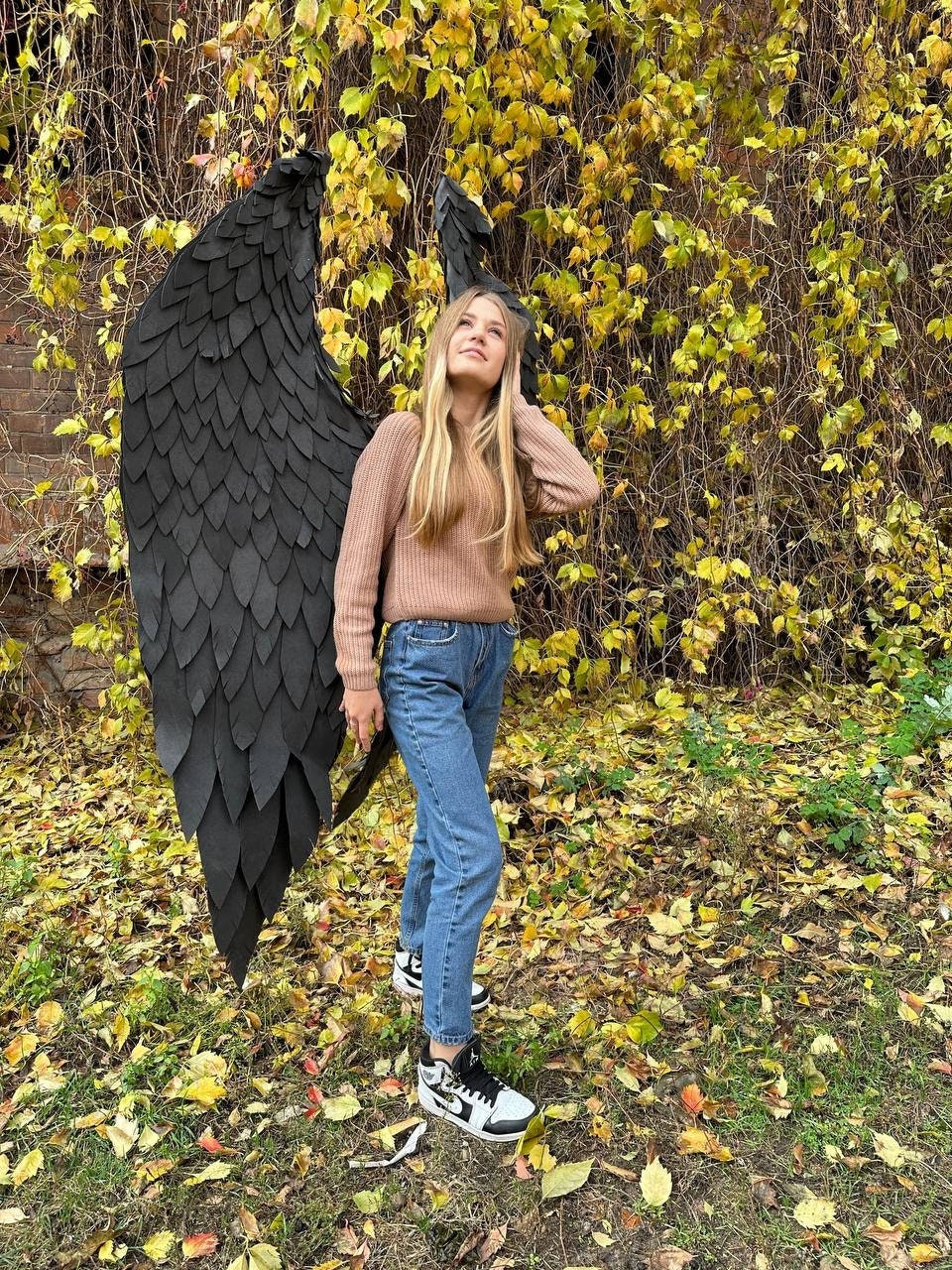 May include: A person wearing a brown sweater and blue jeans is holding large black wings made of feathers. The person is standing in a field of fallen autumn leaves.