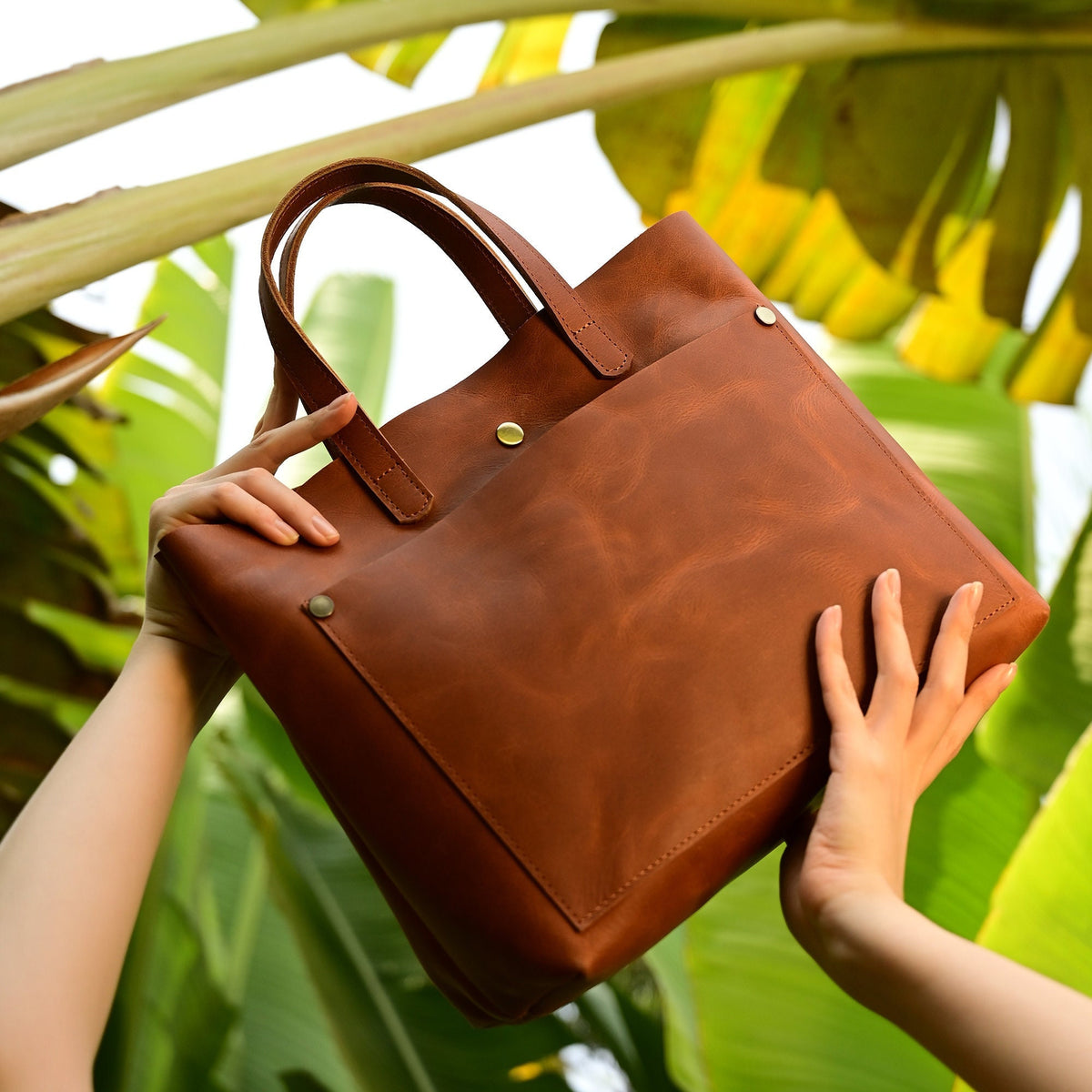 May include: A brown leather tote bag with a large front pocket and a single gold button closure. The bag has two top handles.