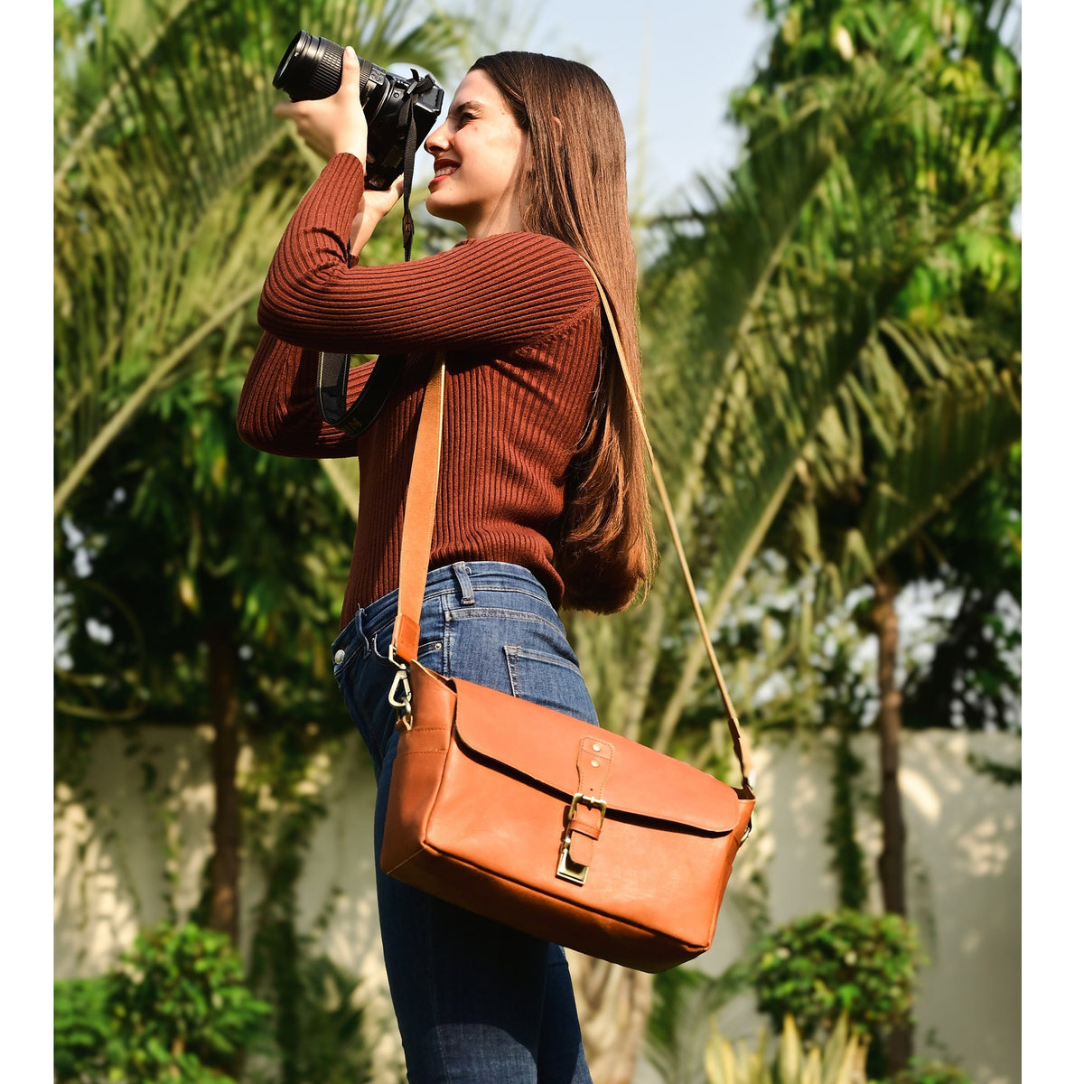 May include: A brown leather camera bag with a shoulder strap. The bag has a rectangular shape with a flap closure and a metal buckle. The bag is worn over the shoulder.