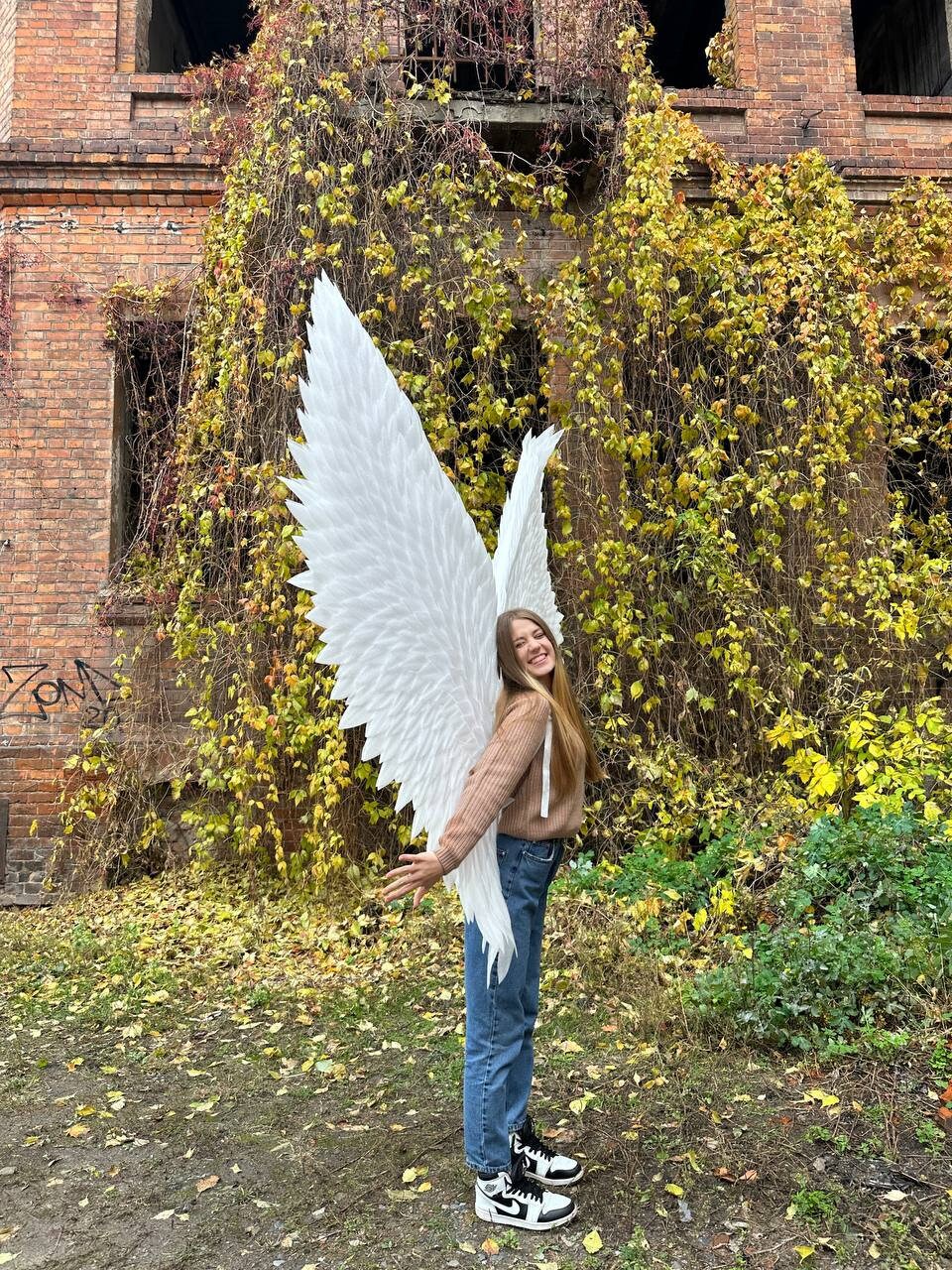 May include: A person wearing a brown sweater and blue jeans is holding large white angel wings in front of a brick building covered in ivy.